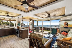 A spacious living area with a large window overlooking the ocean, a kitchen with dark wooden cabinets, and ceiling fans. - Kona Reef#A1: Sand & Surf Beachfront Corner, No Stairs (Kailua Kona)