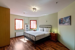 Une chambre à coucher avec un lit, deux fenêtres avec des rideaux, un radiateur, une table de chevet, une lampe et un tableau au mur.