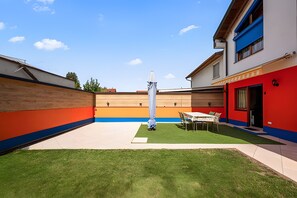 A modern outdoor patio area with a table and chairs, a grassy lawn, and a building with a red and blue facade.
