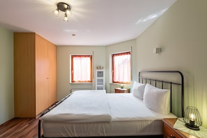 Une chambre à coucher comprenant un lit, une armoire, deux fenêtres avec des rideaux, une table de chevet avec une lampe et un luminaire au plafond.