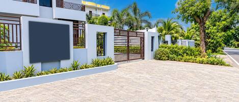 A modern residential building with a paved driveway, a gated entrance, and lush greenery.