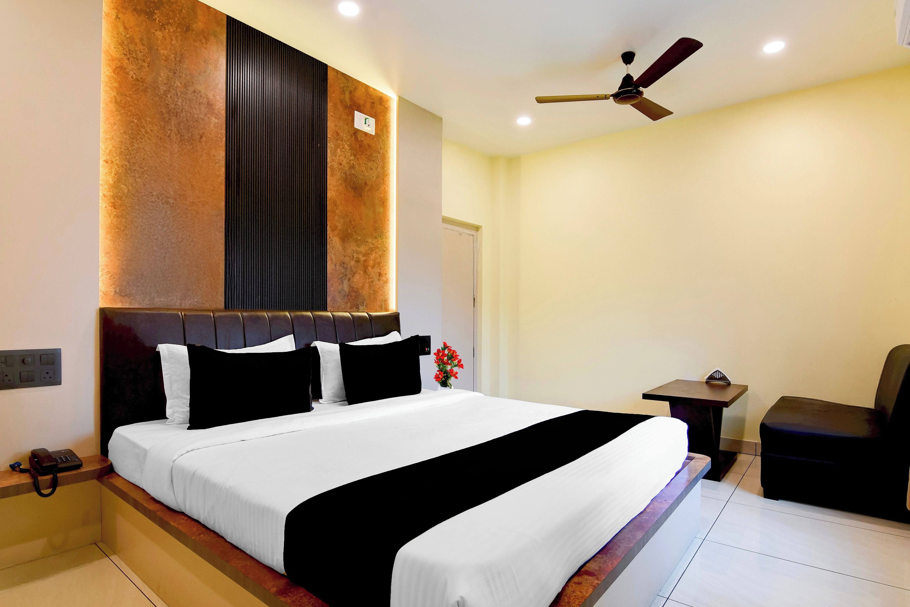 Habitación de hotel con una cama grande, una mesita de noche, una silla, un ventilador de techo y una lámpara fijada a la pared.