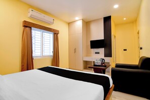 A hotel room with a bed, a desk with a kettle, a chair, a TV, and an air conditioning unit.