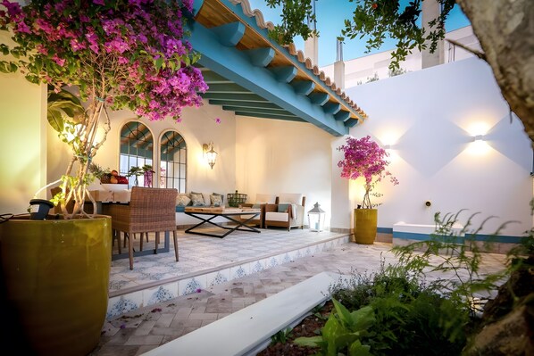 A cozy outdoor seating area with a table and chairs, surrounded by potted plants and a white wall.