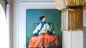 Una habitación con un cuadro grande de una mujer en un vestido azul y rojo, un aparador de madera con dos plantas en macetas, y una lámpara de araña colgante.