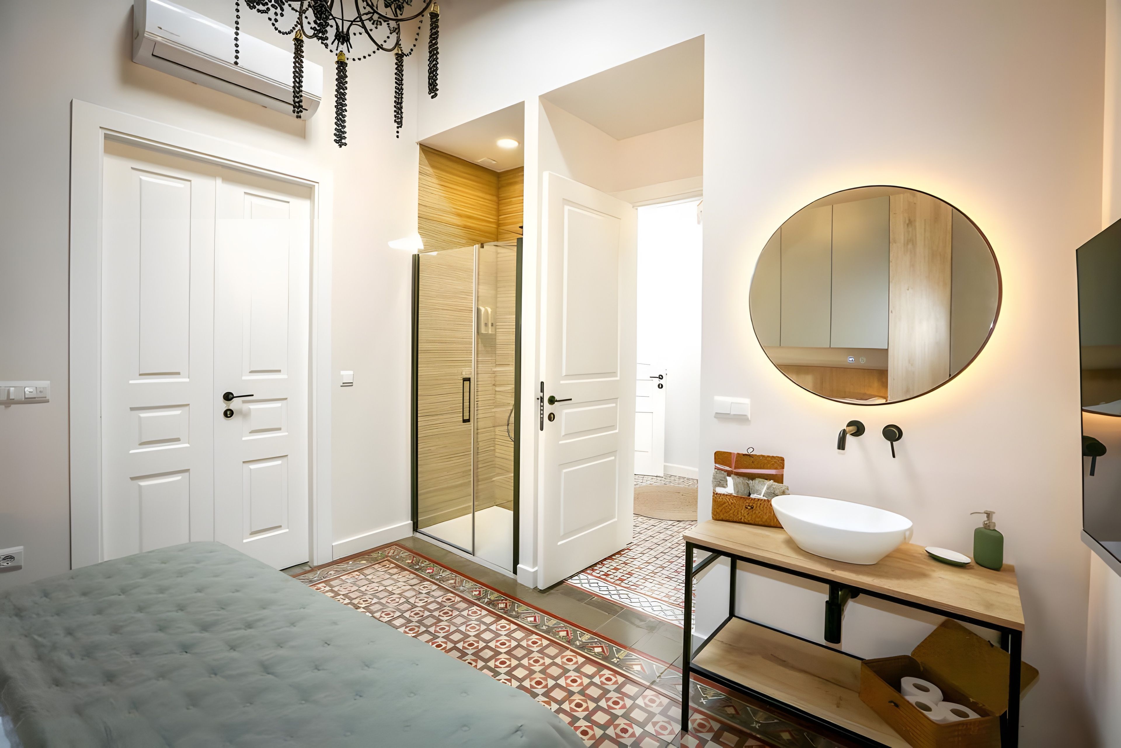Une chambre à coucher moderne avec un grand lit, un dressing, une salle de bain dotée d’un grand miroir rond et d’un lavabo sur colonne, et un sol à motifs.