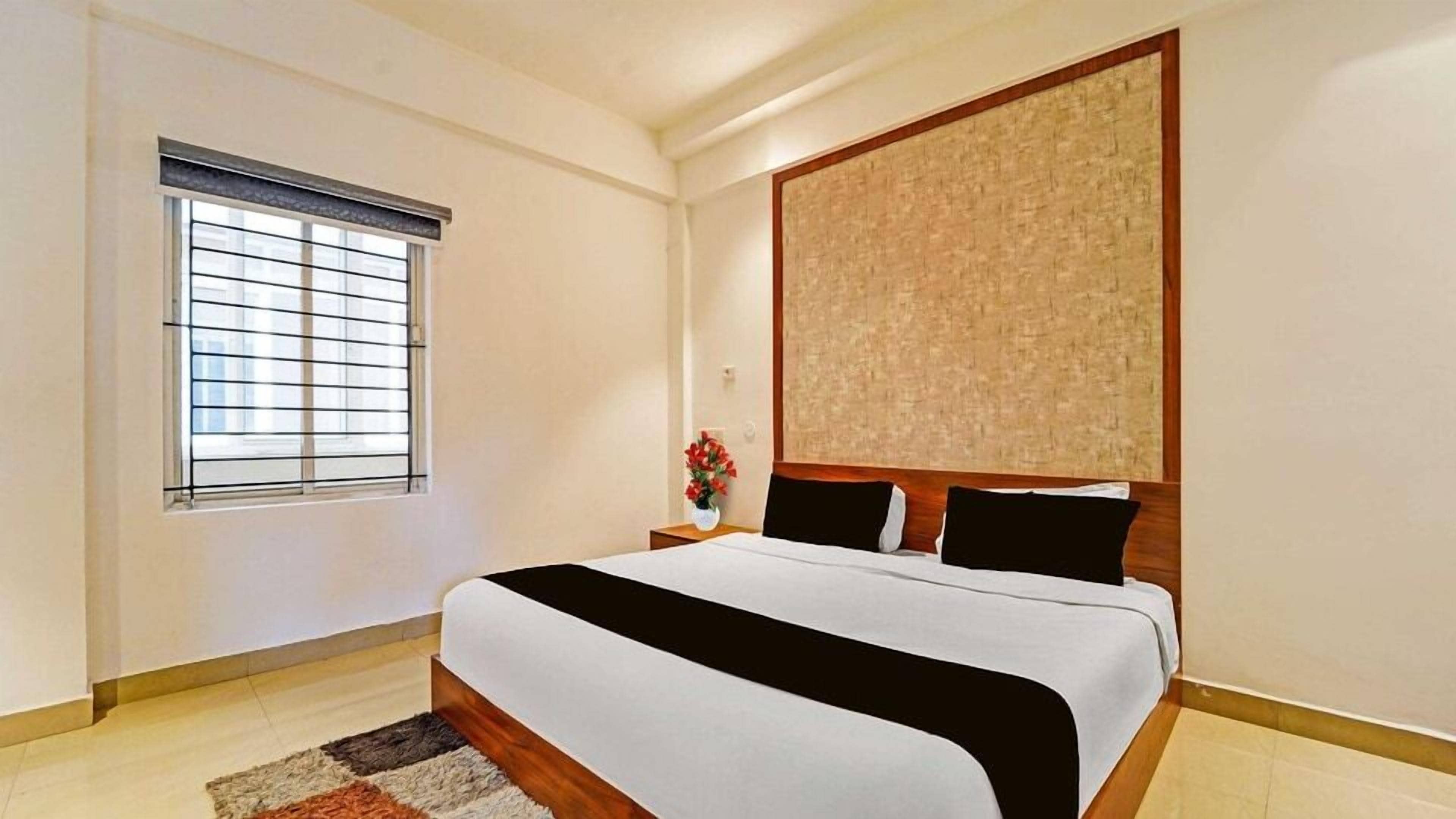 Un dormitorio con cama, ventana con persianas, mesita de noche con un florero y una obra de arte en la pared.