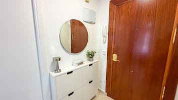 Een gang met een houten deur, een spiegel aan de muur, een kleine potplant, een wit kastje met lades en een telefoon aan de muur.