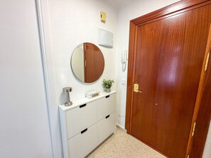 Ein Flur mit einer Holztür, einem weißen Wandspiegel, einem kleinen Topfpflanzen, einem weißen Schrank mit Schubladen und einem Wandtelefon.