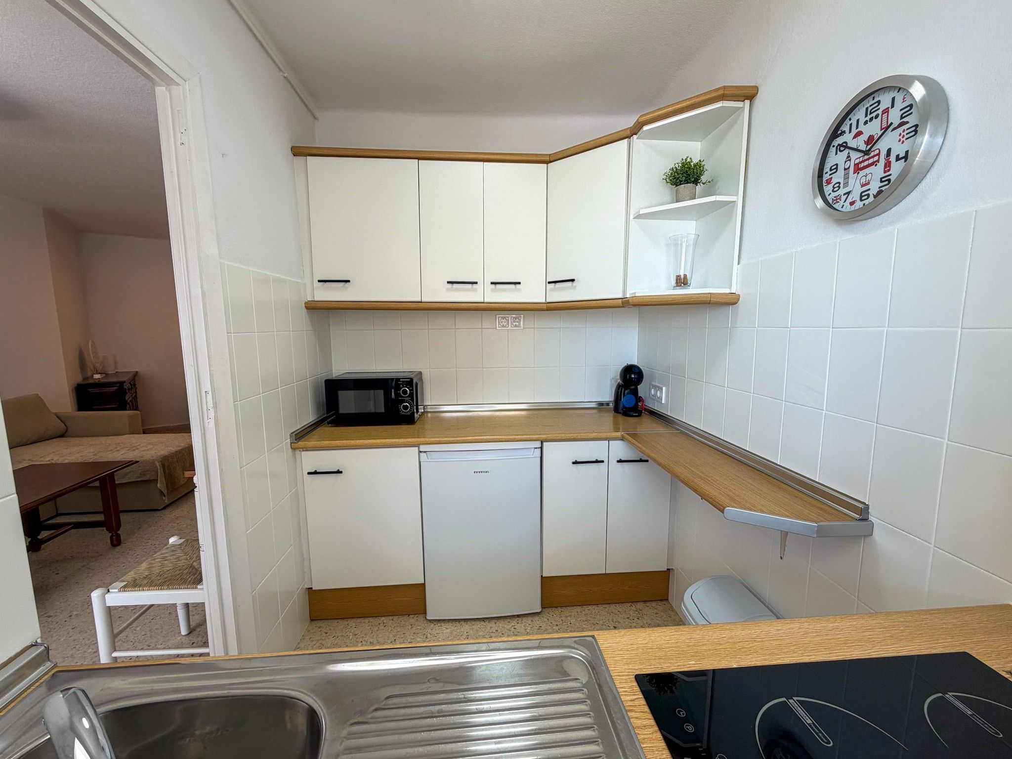 Een kleine keuken met witte kasten, een magnetron, een koffiezetapparaat en een klok aan de muur.