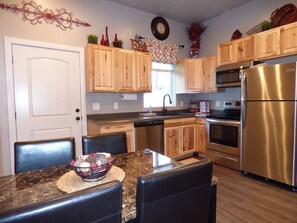 A kitchen with wooden cabinets, stainless steel appliances, and a dining area with a marble countertop and chairs. - Downtown Kanab Hideaway | Walk to Shops & Dining (Kanab)