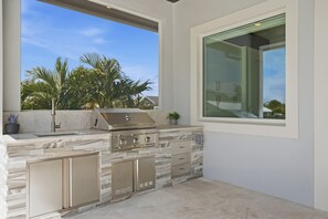 An outdoor kitchen with a built-in grill, a sink, and a window overlooking a residential area. - Luxury Home - Close to Beach -Saltwater Pool & Spa (Naples)
