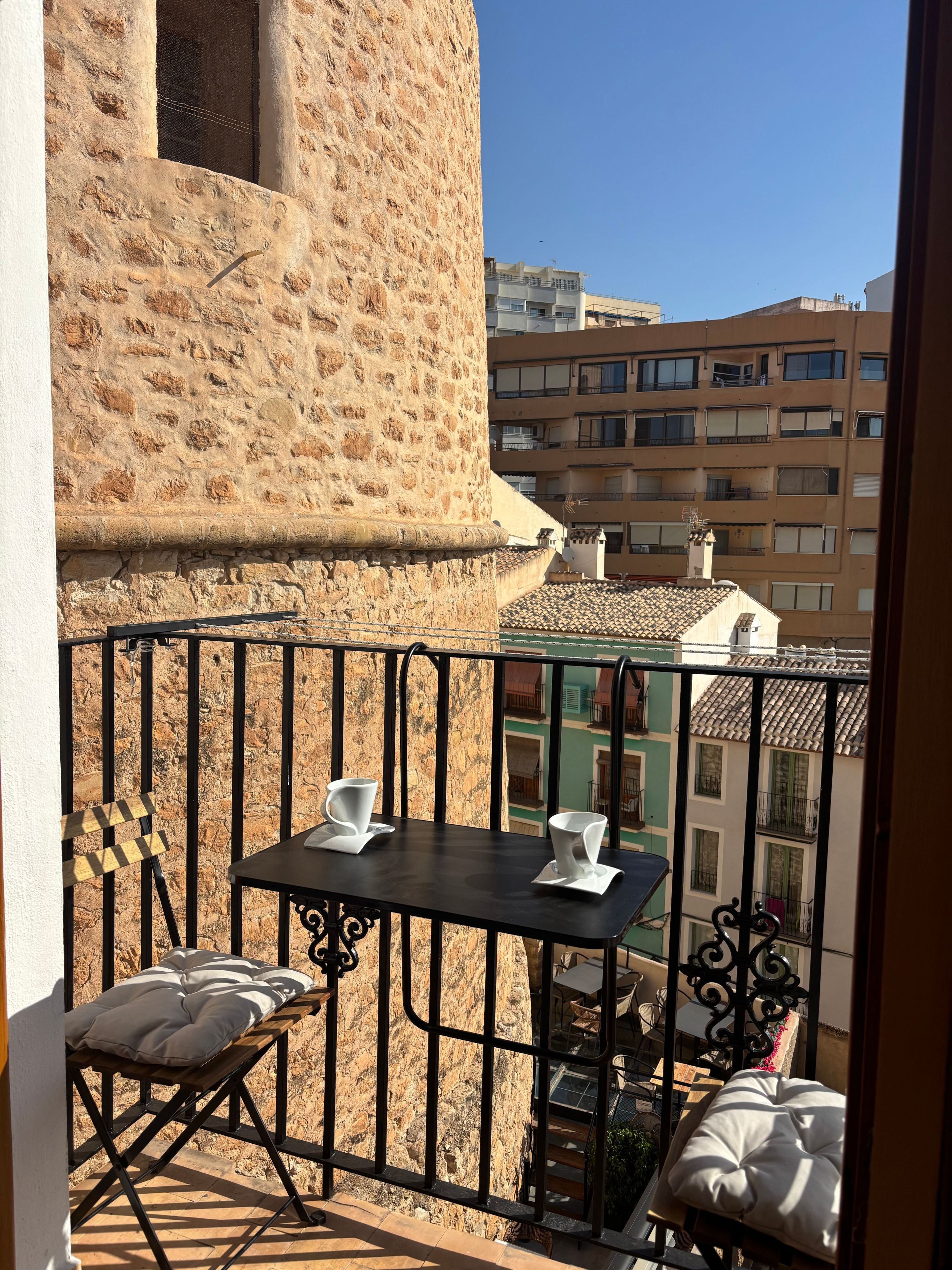 A balcony with a stone wall, a table with two chairs, and a view of neighboring buildings.