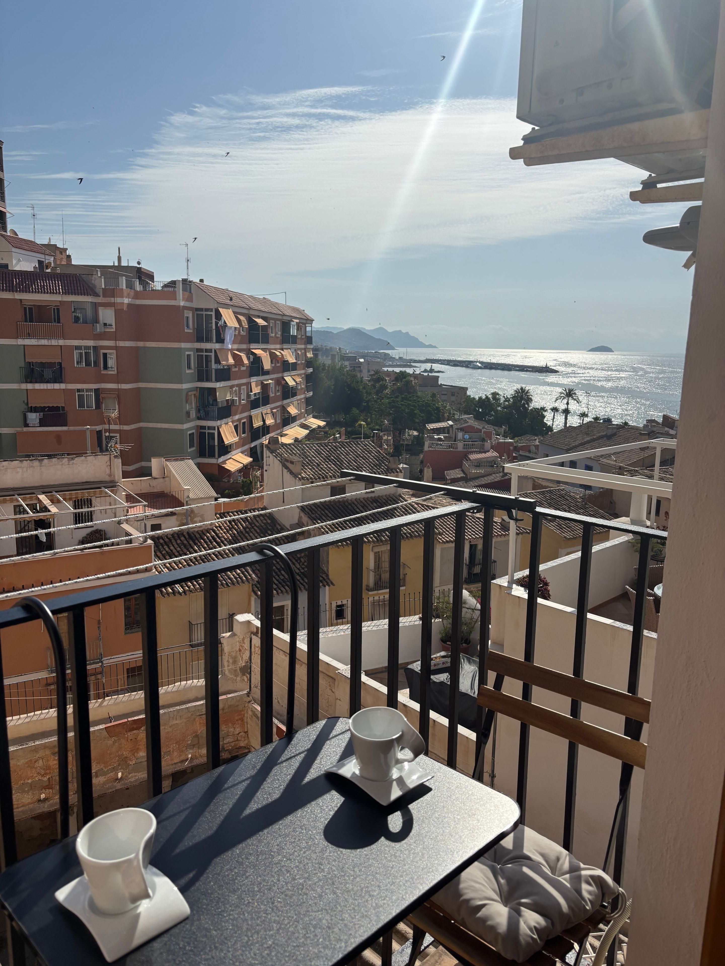 A balcony with a table, a cup, and a view of the sea and buildings.