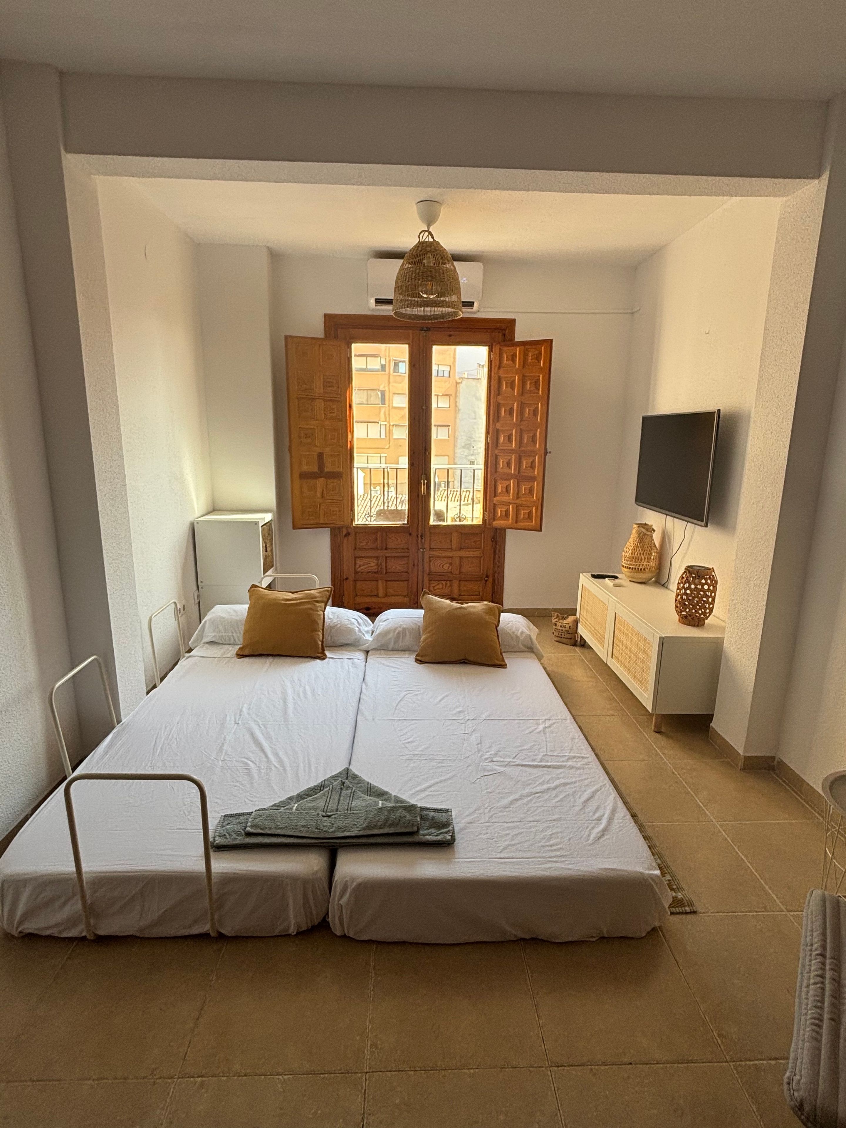 Un dormitorio con cama, televisión, mesita de noche y ventana con contraventanas abiertas.