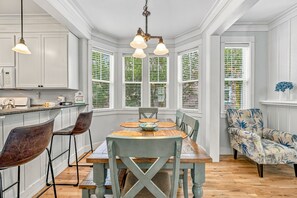 Una cocina con armarios blancos, una mesa de comedor de madera con sillas y un ventanal con follaje verde.