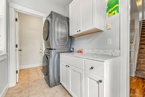 A compact laundry room with a front-loading washer and dryer, a marble countertop, and a small storage cabinet. - Olivia Beach Beauty; Hot Tub, Grill, Near Beach Access (Lincoln City)