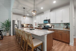 A modern kitchen with white cabinets, a central island with chairs, stainless steel appliances, and a hanging light fixture.