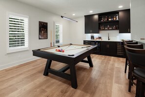 A pool table in a room with wooden flooring, a kitchen area with dark cabinets, and a window with blinds.