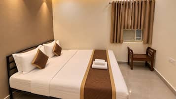 A hotel room with a bed, a bedside table, a chair, a window with curtains, and an air conditioning unit.