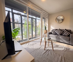 A modern living room with a large window, a grey sofa, a round mirror, and a glass coffee table. - Edificio Quinta Vista by Domus Travels (Quinta Normal)