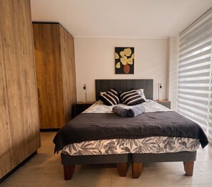 A bedroom with a bed, a nightstand, a lamp, a wardrobe, and a window with blinds. - Edificio Quinta Vista by Domus Travels (Quinta Normal)