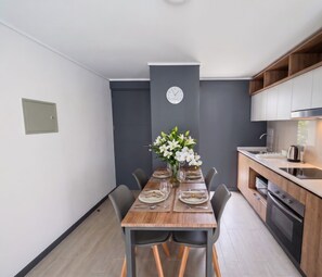 Une cuisine moderne avec un four encastré, une table à manger pour quatre personnes agrémentée d’un vase de fleurs et une horloge au mur.
