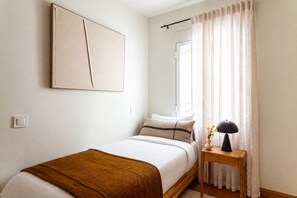 A bedroom with a bed, a bedside table with a lamp, a window with curtains, and a wall-mounted artwork. - A two-bedroom in Chamberí where full-length windows open onto a balcony view and a large round table seats friends for a long meal, on a street where families have lived for generations. (MADRID)