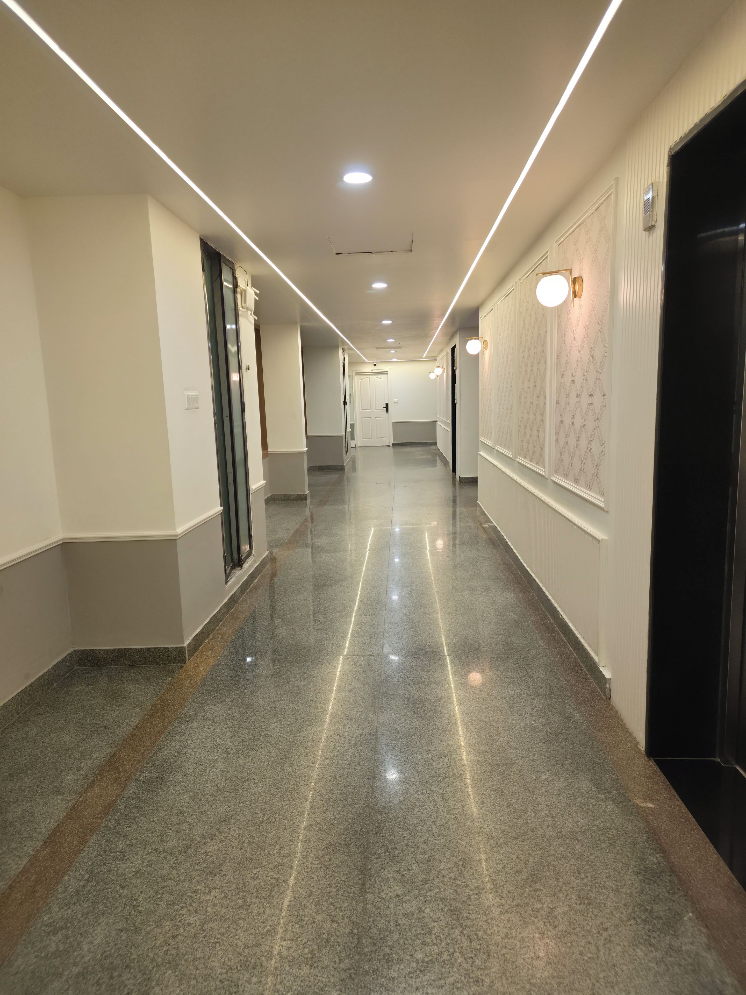 Un pasillo largo y bien iluminado con suelo pulido, puertas a ambos lados y luces empotradas en el techo.