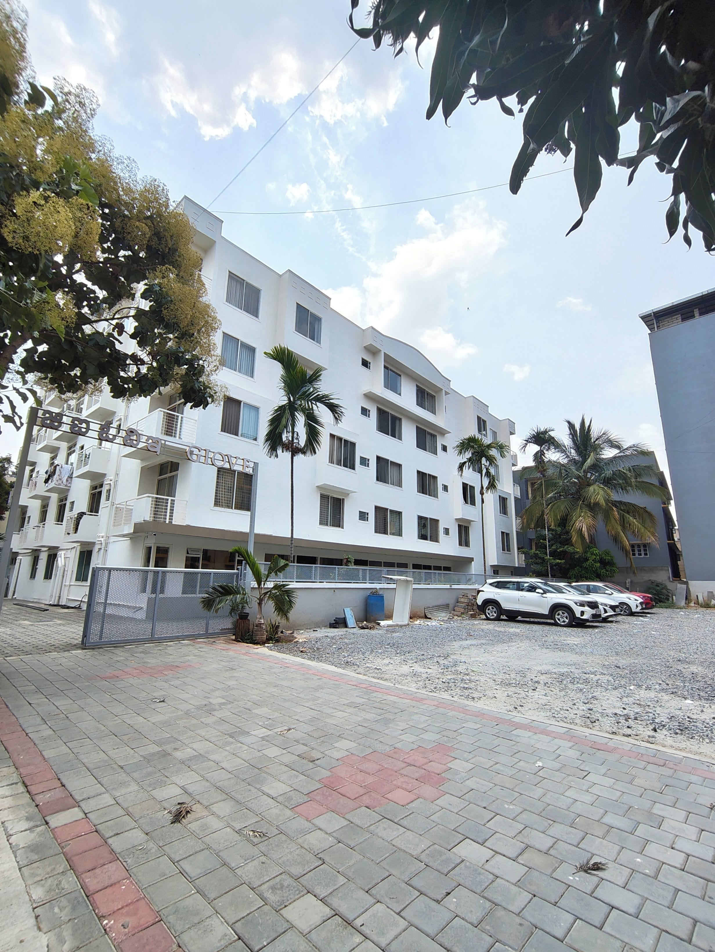 Un edificio de varios pisos con balcones, un área pavimentada con una cerca y coches aparcados.