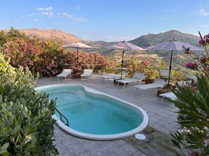 Ein Poolbereich mit Liegestühlen und Sonnenschirmen, umgeben von blühenden Pflanzen und Bergen im Hintergrund.