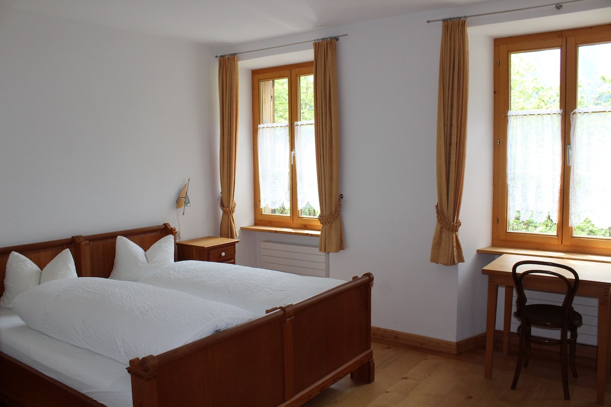 A bedroom with a wooden bed, a desk, and a chair. There are two windows with curtains.