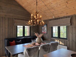 Ein Interieur einer Holzhütte mit Holzbalkendecke, einem Chandelier, einem Sofa, einem Esstisch und einem Fenster mit Blick.