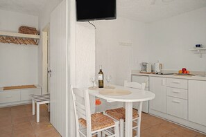 A small, white-walled room with a dining table, chairs, a television mounted on the wall, and a kitchen area with a coffee maker and fruit. - Apartment 'Pool Suite 9' with Shared Pool and Balcony (Αρκάσα)