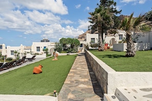 A courtyard with a stone pathway, outdoor seating, and a view of white buildings with blue doors. - Studio Apartment 'Thalassa Suite 3' with Sea View and Shared Pool (Αρκάσα)