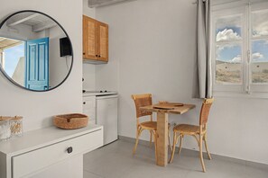 A compact kitchen with a white countertop, a wooden cabinet, a round mirror, and a dining area with two wicker chairs and a small table. - Studio Apartment 'Thalassa Suite 6' with Sea View and Shared Pool (Αρκάσα)