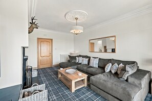 Ein Wohnzimmer mit grauem Sofa, einem Holztisch, einem karierten Teppich, einem Kamin und einem Spiegel an der Wand.