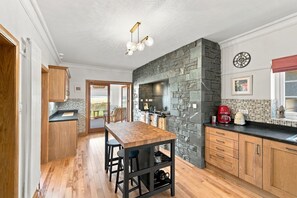 Una cocina moderna con protector mural de piedra, armarios de madera, una mesa de comedor con taburetes y vistas a través de una puerta abierta.