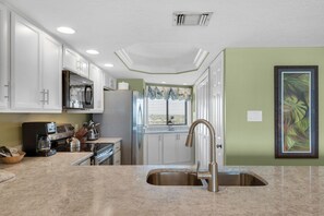 A kitchen with white cabinets, a stainless steel refrigerator, a coffee maker, and a window with blinds. - Edgewater Beach Condo 807 (Destin)