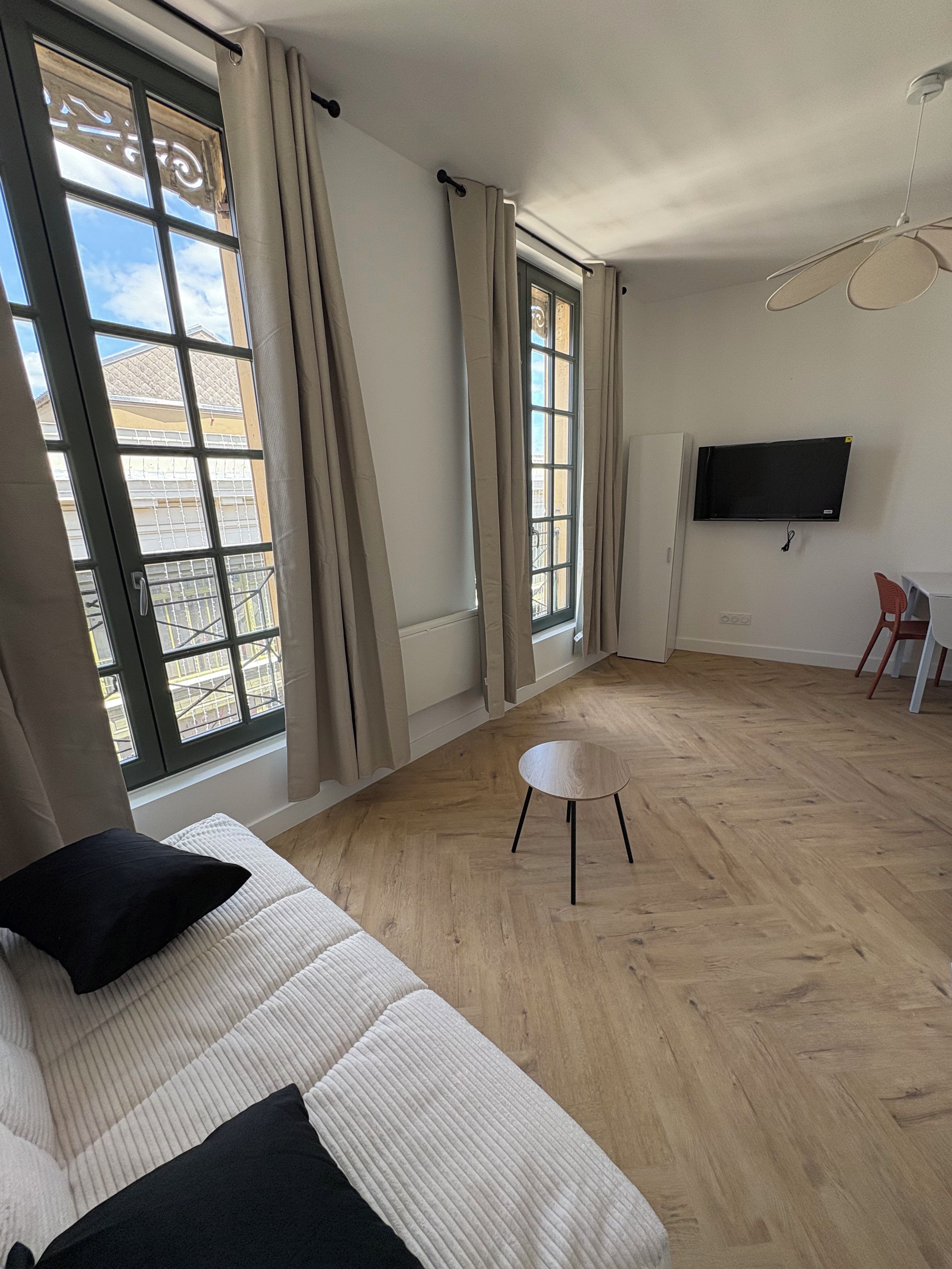 A living room with a sofa, a small table, a TV mounted on the wall, and large windows with curtains.