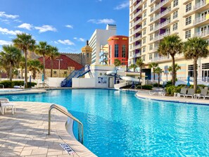 A swimming pool with a water slide, surrounded by palm trees and modern buildings. - Spacious condo Ocean Walk Resort with lazy river (Daytona Beach)