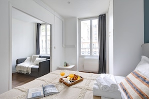 A hotel room with a bed, a sofa, a small table with a tray of breakfast, and a window with curtains. - Cozy 1-Bedroom near Bastille - Nation (Paris)