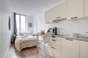 Une pièce moderne et bien éclairée, comprenant un lit, un coin repas avec une table et des chaises, une cuisine avec des meubles blancs et une fenêtre avec des rideaux.