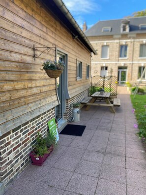 Ein Holzgebäude mit einer Ziegelbasis, einem hängenden Blumenkorb, einem grünen Schild und einem gepflasterten Weg, der zu einem Tisch und einer Bank führt.