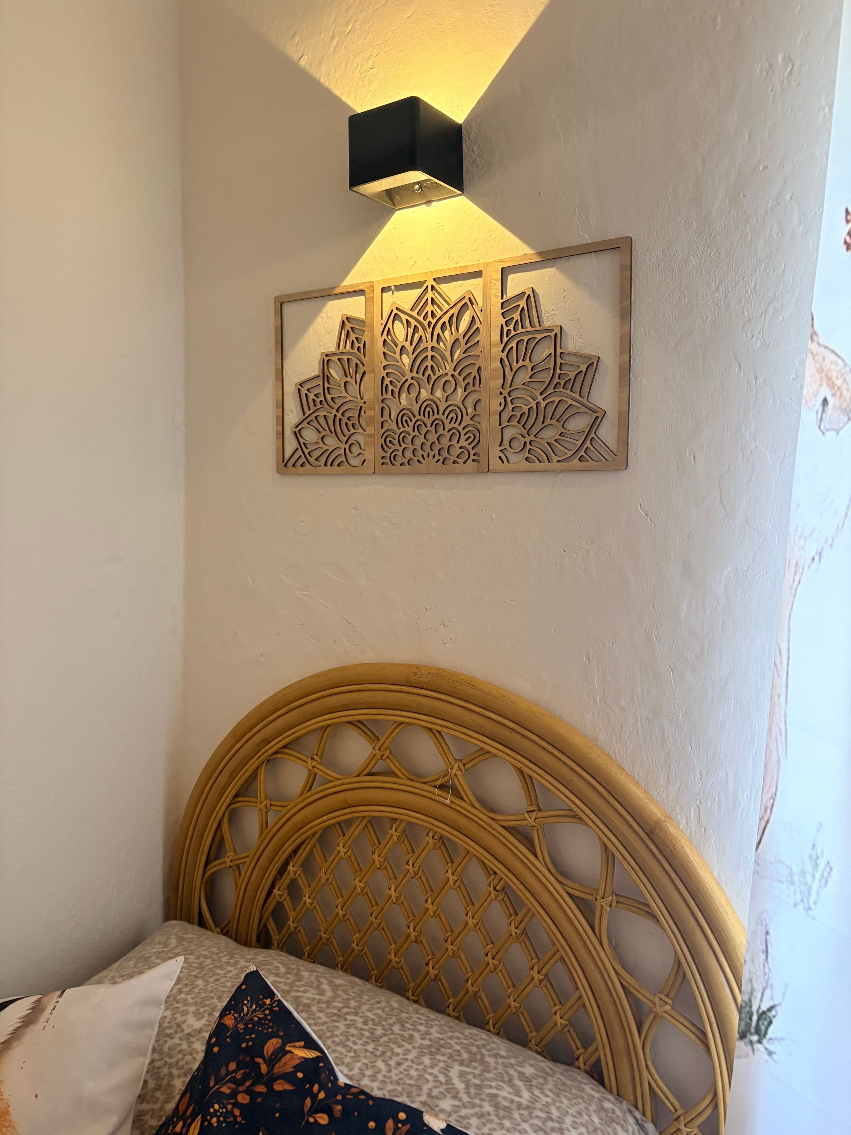 Un lit doté d’une tête de lit en osier, d’un luminaire fixé au mur et d’un panneau décoratif orné de motifs complexes.