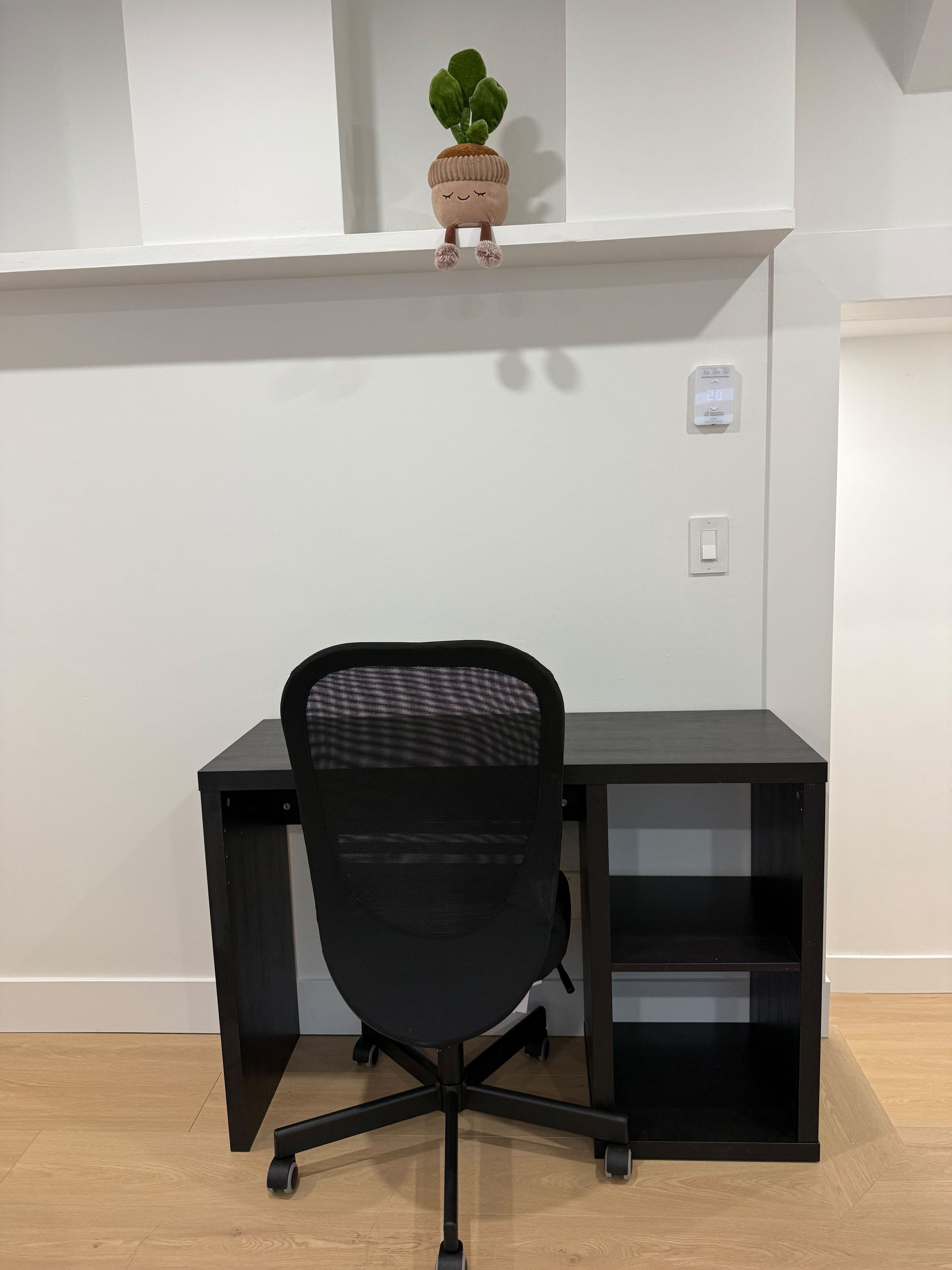 A black office chair with a mesh backrest and wheels, positioned in front of a black desk with a shelf above it.