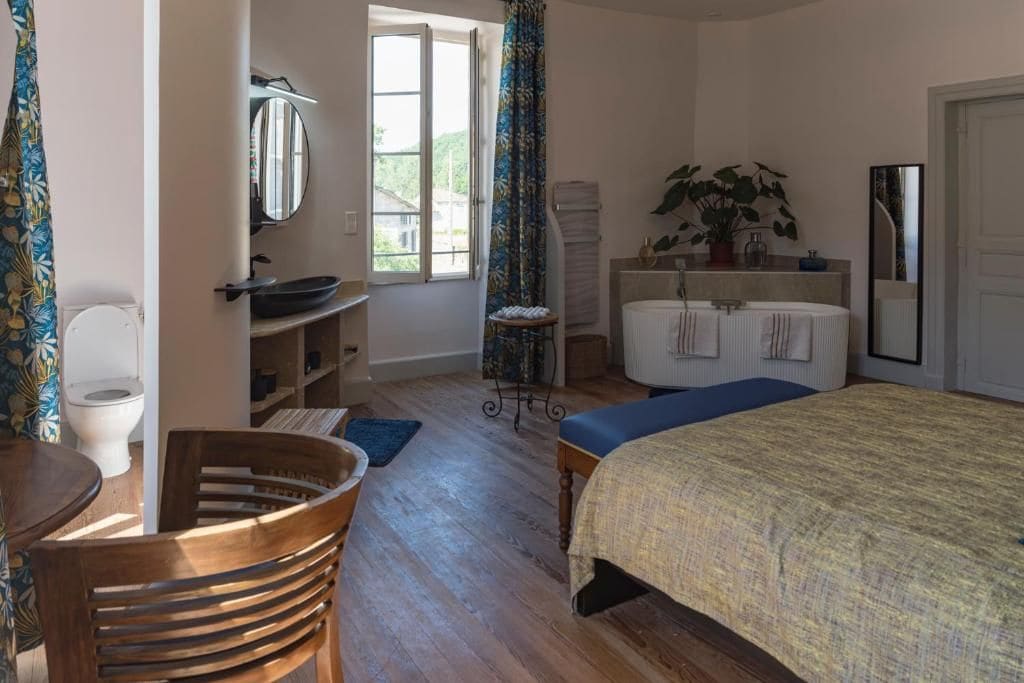 A bedroom with a bed, a bedside table, a chair, a window with curtains, a bathroom with a sink and mirror, and a toilet.