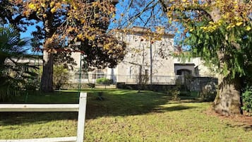 A grassy area with a white fence, trees with autumn leaves, and a building with a stone facade in the background.