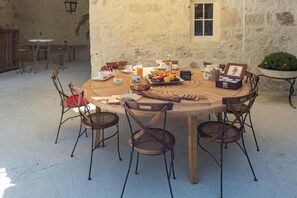 Mesa de madeira redonda com seis cadeiras, rodeada por um muro de pedra, com uma disposição para café da manhã incluindo frutas, pão e bebidas.