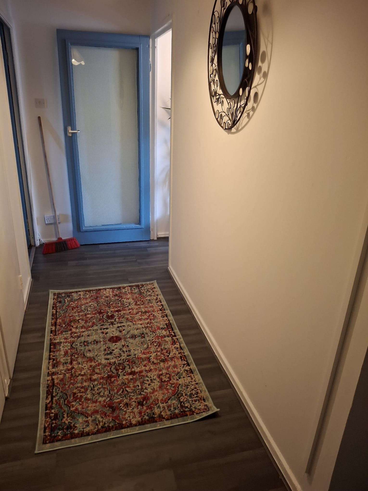 A hallway with a patterned rug, a door with a blue frame, and a wall-mounted mirror.